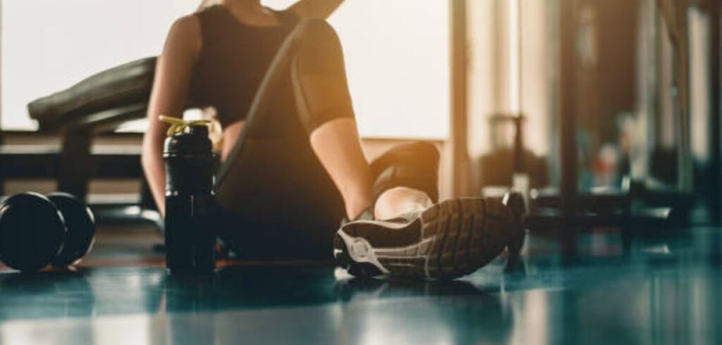 A woman taking a break after doing workout with a protein and dumbbell placed beside at gym. Inside out health and beauty.